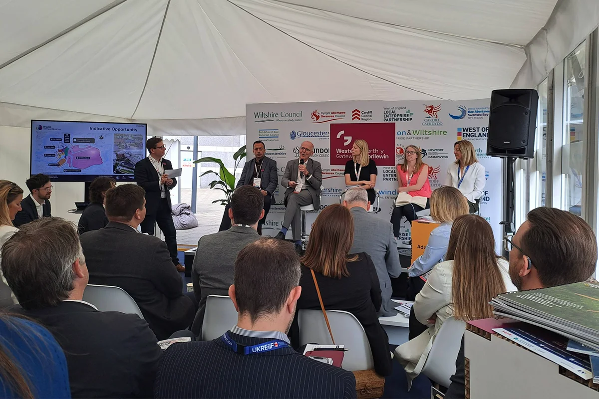 Seated guests with their back to the camera listen to a panel discussion. Richard Bonner, Chair of the West of England Local Enterprise Partnership is standing on the left. Seated from left-to right the panel is Richard Ennis, CEO of the West of England Combined Authority, Stephen Peacock, CEO Bristol City Council, Sophie Allan, Director at Deloitte, Marianne Brooke, Director at Homes England, and Kirsten Durie, Industry Programme Manager for Network Rail.