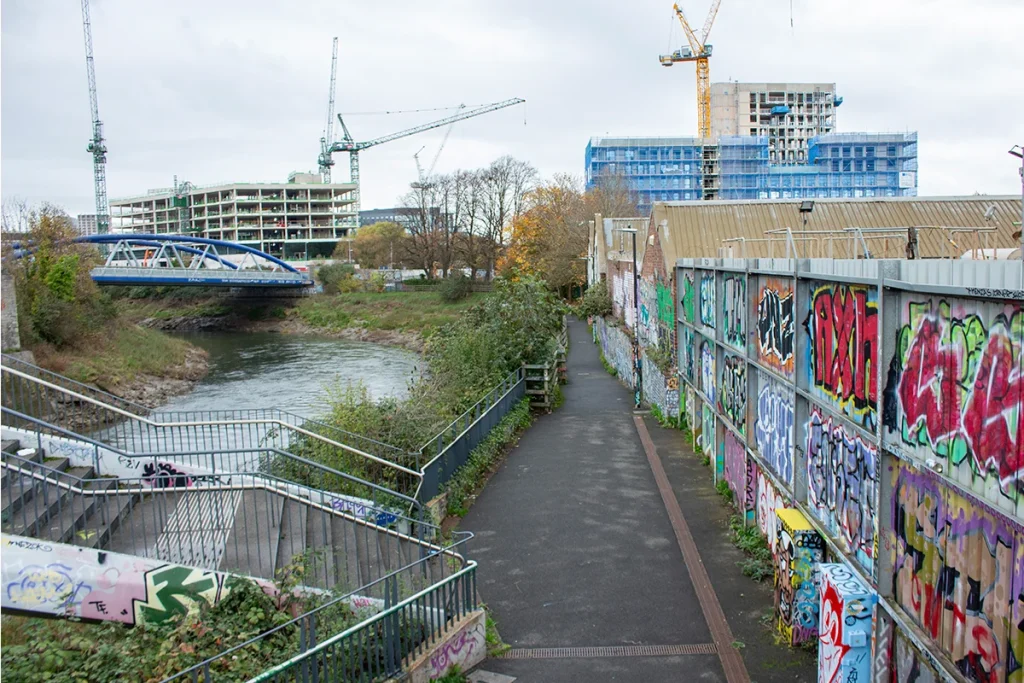 - Photo of the River Avon walking and cycling path. The river is on the left. On the right there are warehouses and a wall with graffiti on it.